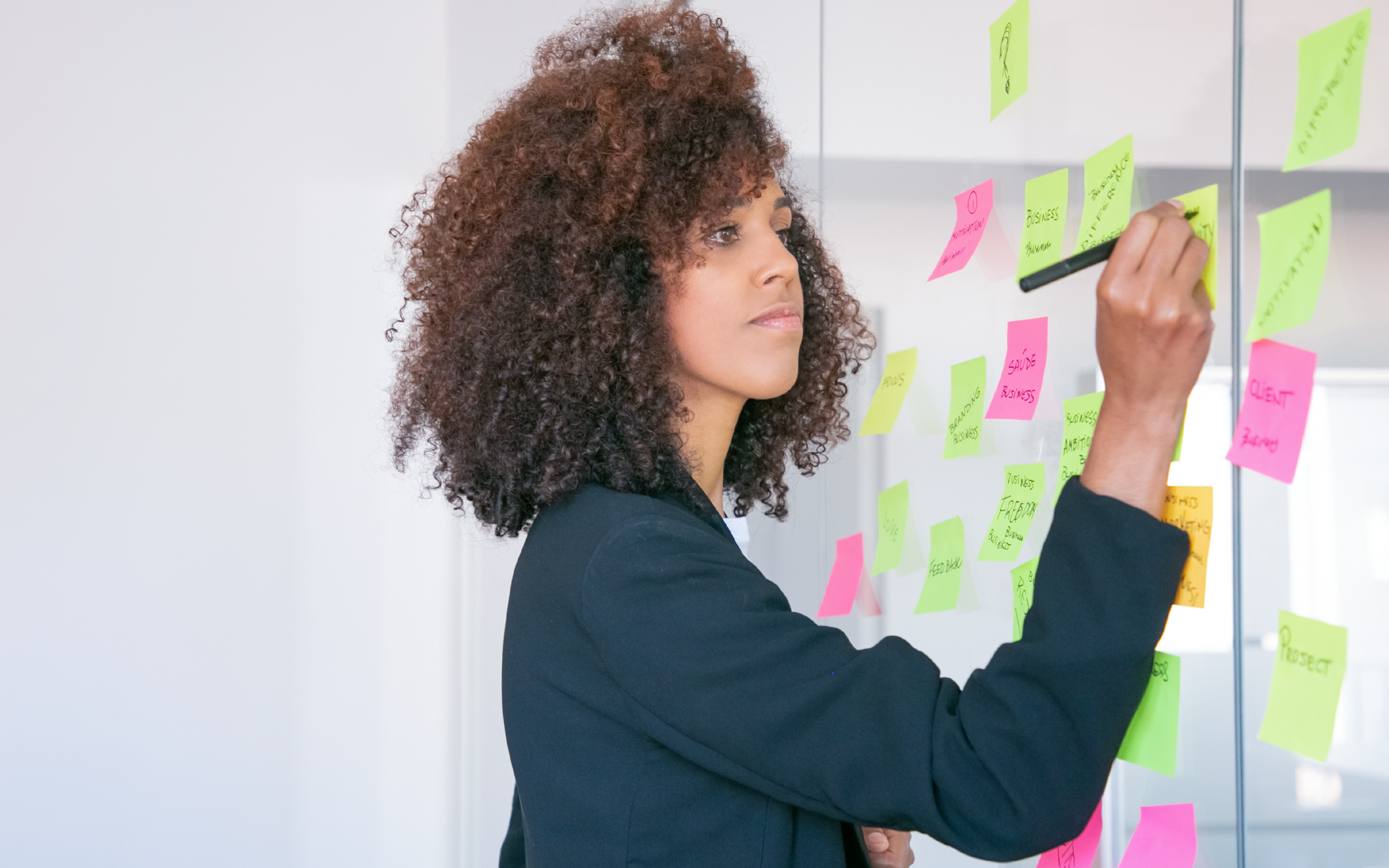 Mulher negra escrevendo em post its coloridos que estão fixados em um quadro de vidro. Ela planeja nesse mural possíveis novas estratégias para a área de Gente e Gestão da empresa onde trabalha.