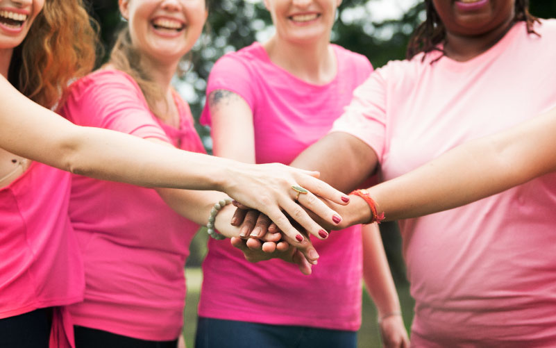 A imagem mostra ums grupos de quatro mulheres utilizando blusa rosa. Elas estão com as mão unidas ao centro, uma sobre a outra. A imagem busca representar a importância da campanha Outubro Rosa nas empresas.