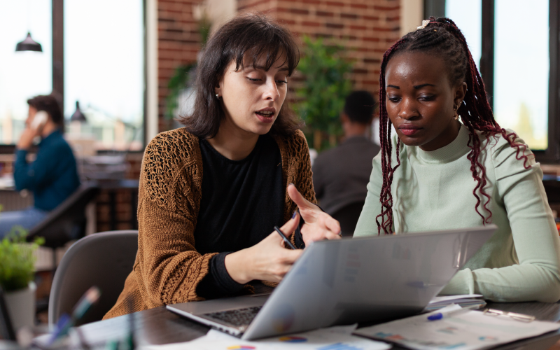 Imagem de duas mulheres sentadas conversando enquanto olhar para a tela de um computador. Ela busca representar a criação de um programa de desenvolvimento de liderança.