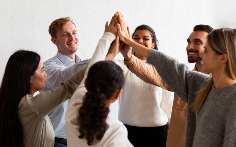 Grupo de profissionais sorrindo enquanto unem aos mãos ao centro, em gesto de comemoração. A imagem busca representar o comportamento organizacional.