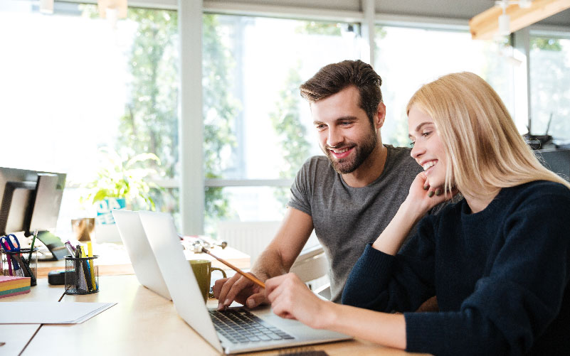 Homem e mulher sentados lado a lado em uma mesa de escritório. Eles analisam informações enquanto observam a tela de um notebook. A imagem busca representar a estruturação das etapas do funil de recrutamento e seleção.