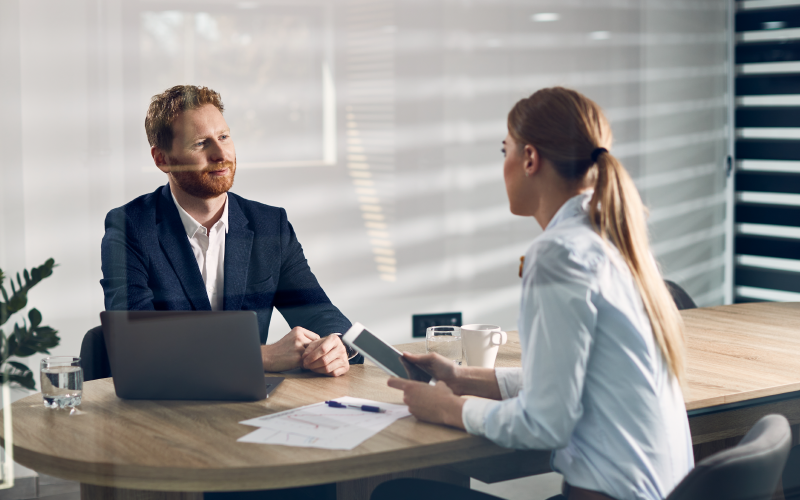 Um homem e uma mulher sentados ao redor de uma mesa de escritório, um de frente para o outro. Eles estão conversando e tem perto de si papéis e um computador para realizar anotações. A imagem busca representar uma reunião de one on one.