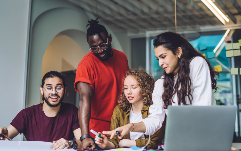 Grupo com quatro jovens dialogando em um ambiente de trabalho. Eles utilizam trajes despojados e observam informações em documentos e em um notebook, que estão apoiados em uma mesa. O objetivo da imagem é representar a atuação de uma equipe multidisciplinar.