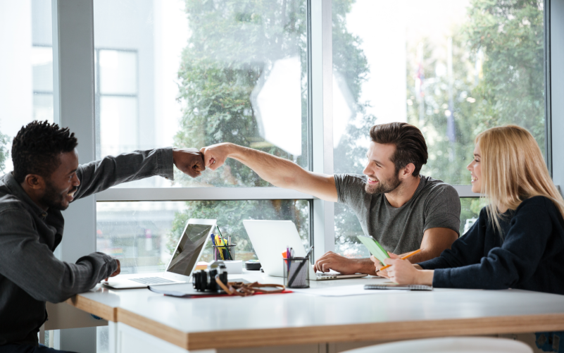 Três profissionais sentados ao redor de uma mesa, em um espaço de trabalho. Os dois homens se cumprimentam em comemoração e a mulher sorri os observando. O objetivo da imagem é demonstrar os benefícios de uma liderança humanizada para empresa e colaboradores.