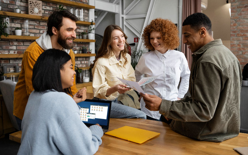 Grupo de pessoas conversando descontraídas ao redor de uma mesa de madeira. O objetivo da imagem é representar a construção de estratégias de team building.