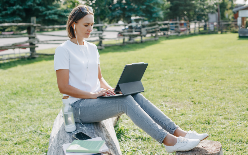 Mulher sentada ao ar livre, com trajes despojados e com um notebook apoiado nas pernas. A imagem busca representar o anywhere office na prática.