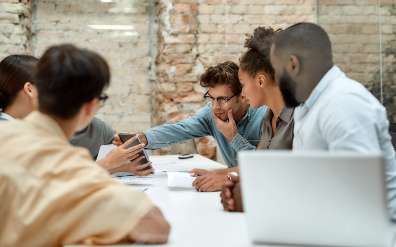 Sala de reuniões com mesa ao centro, em que grupo de profissionais está sentado e conversa sobre os relatórios apresentados. A imagem busca representar a atuação de high performers.