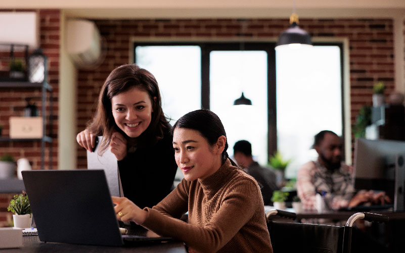 Duas mulheres conversando enquanto olham para a tela de um computador portátil. A imagem busca representar o incentivo ao intraempreendedorismo nas empresas.