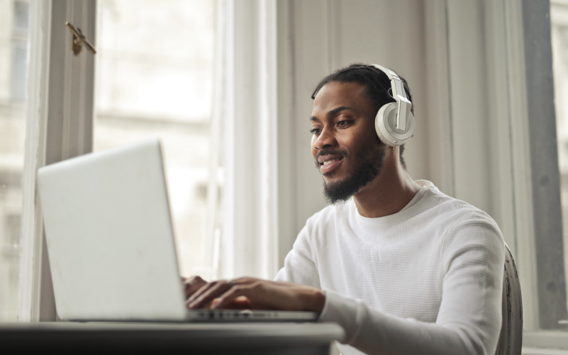 Homem negro utilizando headphones sentado em frente a um notebook. A imagem busca representar a participação em um processo seletivo no metaverso.