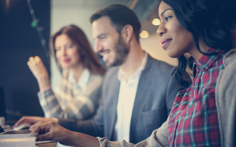 Três profissionais sentados lado a lado olhando para uma tela. Eles tem um semblante facial que representa satisfação e parecem estar observando algo novo. O objetivo da imagem é representar a implementação de um sistema integrado no RH.