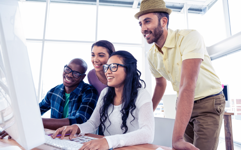 Grupo de quatro pessoas sorrindo, enquanto olham em uma mesma direção. Elas parecem observar uma tela, e uma das mulheres está usando um teclado de computador. A imagem busca representar o uso de um software de OKR.