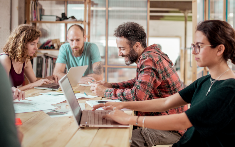 Grupo de pessoas sentadas ao redor de uma mesa de madeira. Elas estão em um ambiente descontraído e conversam observando as telas de seus notebooks. O objetivo da imagem é representar o processo de desenvolvimento com o uso do upskilling.