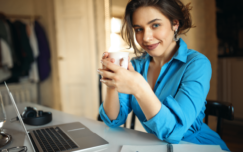 Mulher sorrindo e confiante no trabalho, planejando as melhores formas para alcançar a eficiência operacional na sua equipe. Ela veste uma blusa azul, está segurando uma xícara e está com os braços apoiados em uma mesa de trabalho.