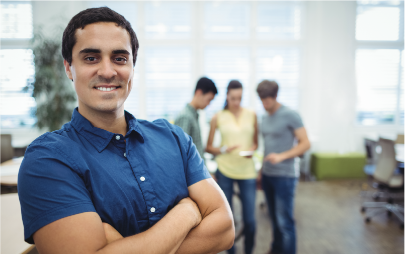 Foco em um homem sorrindo, assumindo a postura e as responsabilidade de ser uma liderança positiva em uma empresa. Ao fundo, três pessoas aparecem desfocadas.