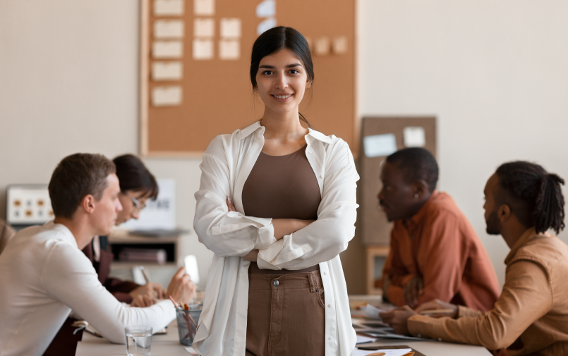 Equipe reunida para treinamentos corporativos na empresa onde trabalham. A imagem traz um grupo de pessoas diversas conversando, e destaca uma mulher que está de pé e sorrindo.