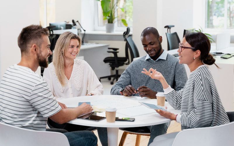 Pessoas diversas - homens e mulheres com diferentes aparências - conversando sentadas a uma mesa de um escritório, representando uma empresa inclusiva
