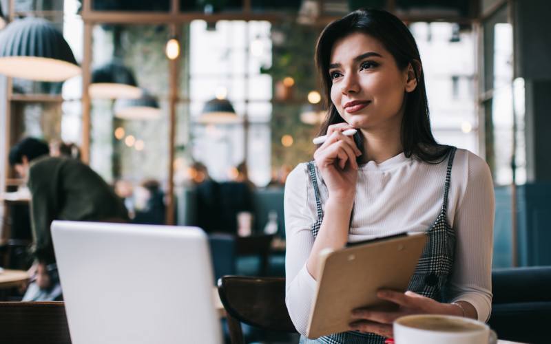 Mulher em ambiente corporativo com olhar reflexivo na frente de um computador, com prancha e caneta em mãos, simbolizando o processo de construção do life design