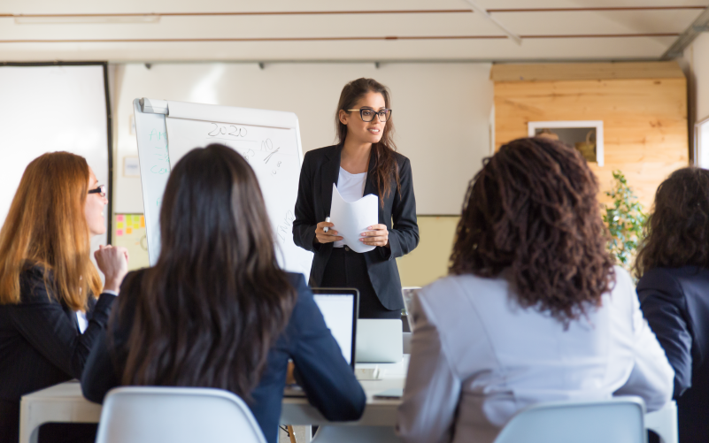 Mulher em pé na frente de outras mulheres sentadas, em posição de liderança inclusiva