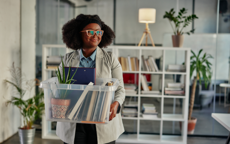 Imagem de capa de um blog post sobre processos de offboarding nas empresas. Na foto, o foco é uma mulher jovem e negra saindo de seu trabalho. Ela carrega seus pertences e está com uma expressão tranquila no rosto.