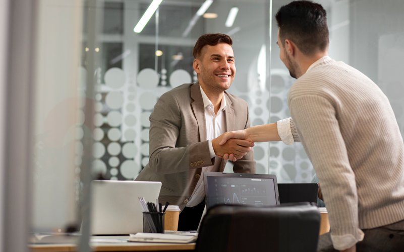 Imagem de capa de post blog sobre promoções internas nas empresas. Na foto, observa-se dois homens jovens. Um deles está sendo cumprimentado pelo outro, dando a ideia de que foi promovido no trabalho.