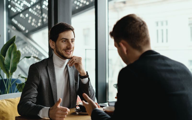 Imagem de post blog sobre interim management. Na foto, observamos dois homens jovens em uma conversa. Ambos estão sentados em uma mesa e um deles aparece na imagem de costas, deixando o foco no outro.