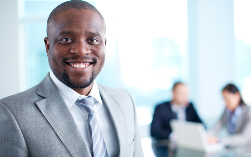 Homem negro sorridente usando terno cinza e gravata, representando um CEO de empresa confiante em ambiente corporativo, com equipe desfocada ao fundo.